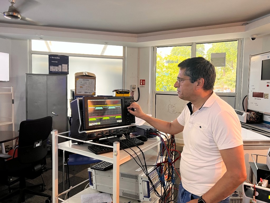 Kunwer Sachdev adjusting a Teledyne LeCroy oscilloscope in an electronics R&D lab, waveforms visible on the screen