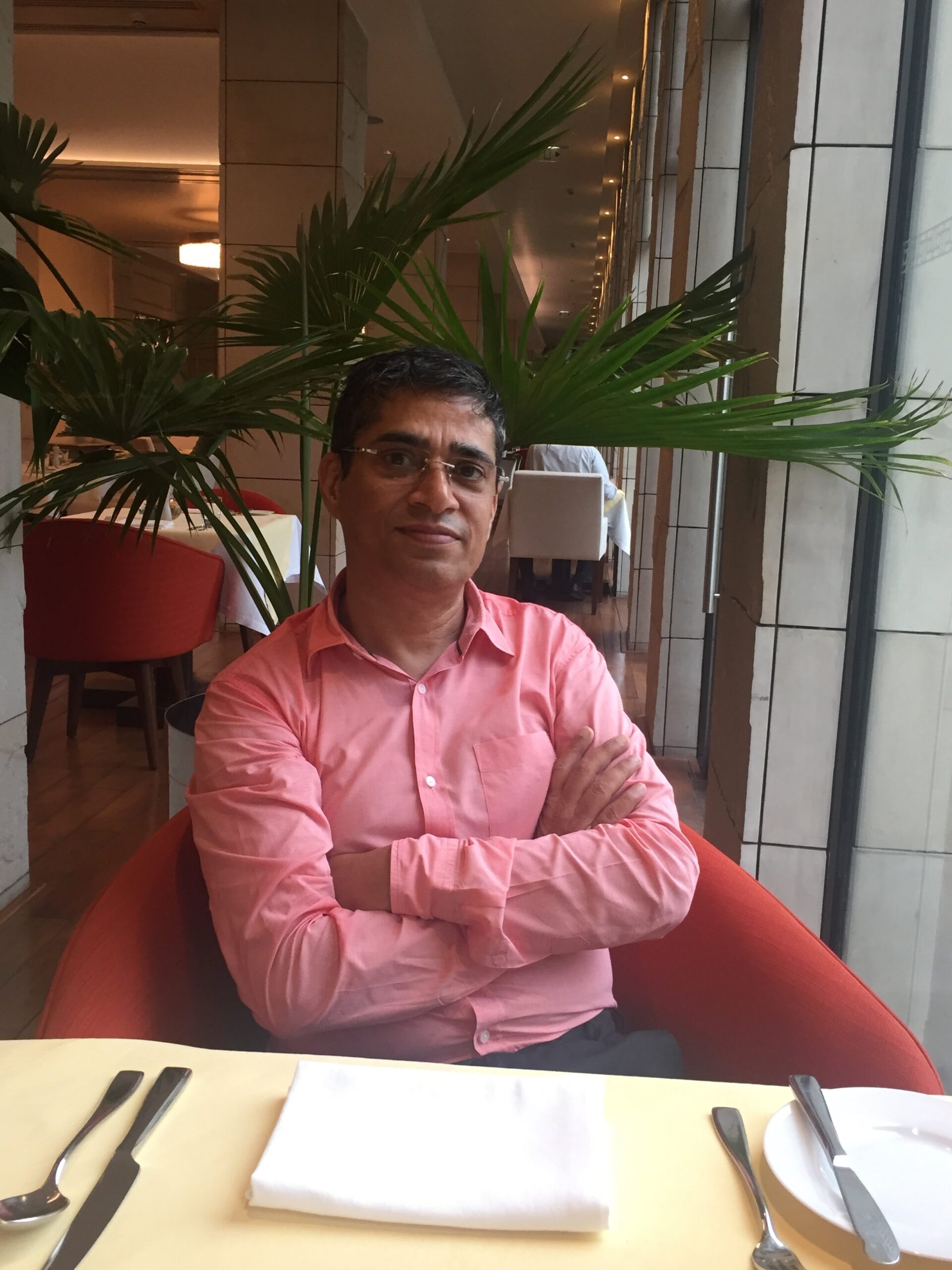 Kunwer Sachdev sitting alone at a restaurant table, arms folded, looking calmly at the camera