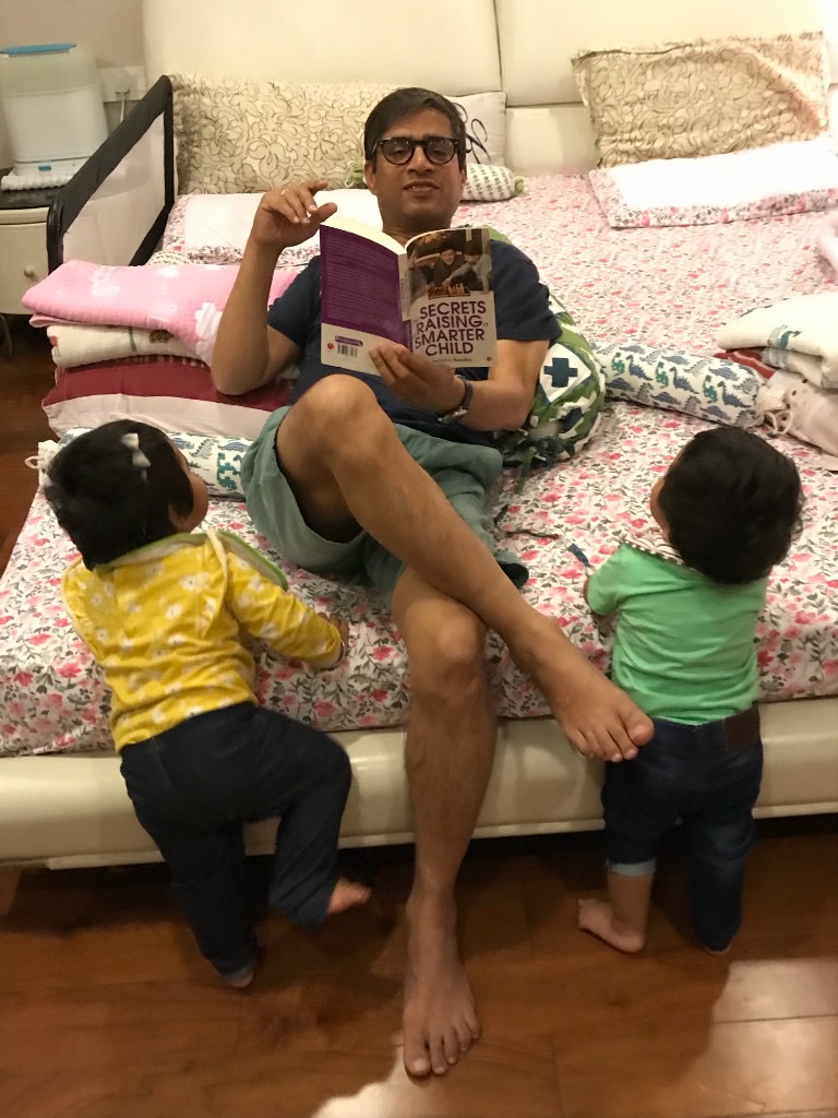 Kunwer Sachdev sitting on a bed reading a parenting book aloud while two young children look up at him
